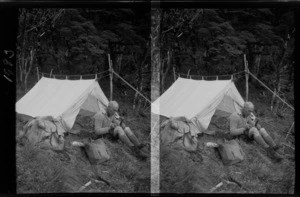 An unidentified man on a mountaineering trip, sitting outside a tent next to backpacks, smoking a pipe and writing in a notebook, location unidentified