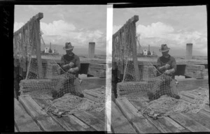 An unidentified man sitting on a wharf smoking a pipe while repairing a fishing net, location unidentified
