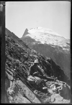 Edgar Williams eating lunch on a mountain ledge, with peak beyond, [Fiordland National Park?]