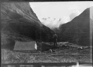 An unidentified man at an alpine campsite, looking at view below partly obscured by cloud, [Fiordland National Park or Canterbury Region]