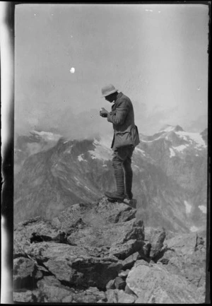 Edgar Williams, on a mountaineering expedition, standing on a rocky outcrop and holding a metal measuring device, with view of mountains below, [Canterbury Region or Southland District?]