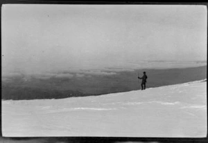 An unidentified young man with a pole, standing on a snowy mountain slope, view of plains below, probably Canterbury Region