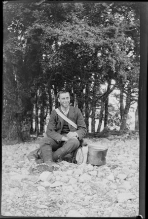 An unidentified young man sitting on a pack, smoking a pipe, during a mountaineering expedition, [Canterbury, Otago or Southland?]