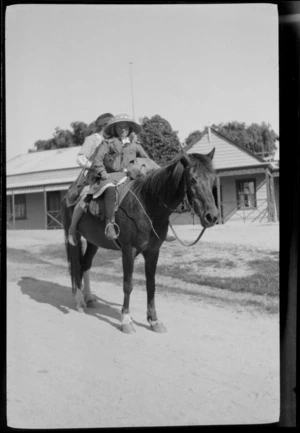 Two unidentified girls, one holding a switch, on the back of a horse on a street with wooden buildings beyond, [Canterbury, Otago or Southland?]