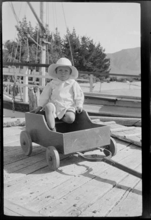 An unidentified child wearing a wide-brimmed hat, sitting in a wooden wagon on wharf, [Lake te Anau], Southland Region
