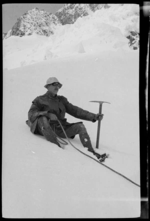 Edgar Williams, wearing an oilskin coat and goggles with rope tied around waist and holding icepick, sitting on a snowy mountain slope during a climbing expedition, [West Coast Region]
