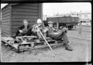 Edgar Williams, centre, with two other men [Adam Gorton and Mr Watkin?], with mountaineering gear, sitting on a railyard platform, carriages beyond, [Ashburton District, Canterbury Region?]