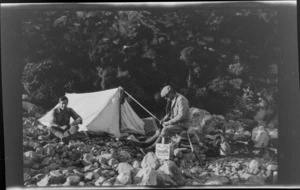 Edgar Williams and an unidentified man [Tracy Thomas Gough?], at a campsite, with rifle, biscuit box and backpacks on stony ground outside tent, Ashburton District, Canterbury Region