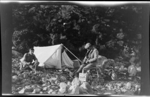 Edgar Williams and an unidentified man [Tracy Thomas Gough?], at a campsite, with a biscuit box, rifle, and backpack spread on stony ground outside tent, Canterbury Region