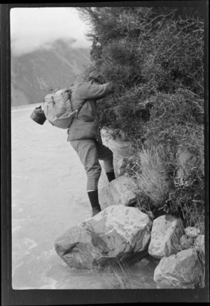 A man [Tracy Thomas Gough?], climbing on boulders next to a river, Southern Alps, Canterbury Region