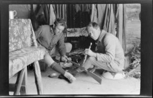 Edgar Williams, holding a flute, and another man [Tracy Thomas Gough?] cleaning a rifle, sitting in front of fireplace in mountain hut, Southern Alps, Canterbury Region