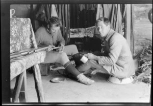 Edgar Williams, right, and another man [Tracy Thomas Gough?], who is preparing food, sitting in front of a fireplace in a mountain hut, Southern Alps, Canterbury Region