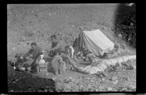 Edgar Williams and an unidentified man at a camp on a moraine, boiling a billy over a gas burner, tent and sleeping bag on stony ground behind, near Lyell Glacier, Southern Alps, Canterbury Region