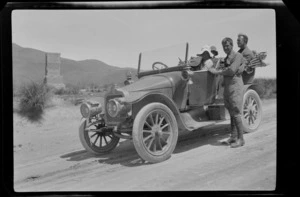 An unidentified group in a motorcar, on an unsealed road next to memorial dedicated to Michael John Burke, Burke Pass, Mackenzie District