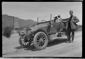 Two unidentified men and a girl, in a motorcar on an unsealed road next to a memorial, Burke Pass, Mackenzie District