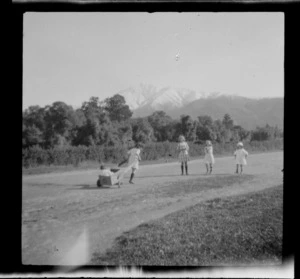 Group of unidentified children on road, one pulling a wooden cart, snow-capped mountains in distance, Peel Forest, Timaru District