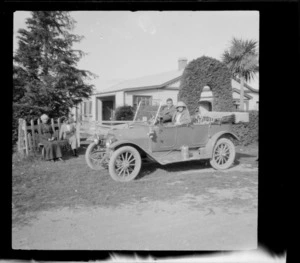 Alice Hazlehurst and Edgar Williams in a motorcar, with two unidentified elderly woman women sitting on a bench nearby, outside a one-storied wooden house with a topiary arch in garden, [Peel Forest, Timaru District?]