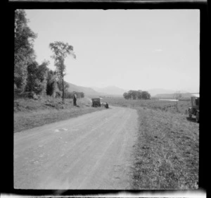 Road through farmland, including two unidentified people standing next to a motorcar [fixing a tyre?], Peel Forest, Timaru District