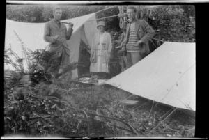 Edgar, Alice and Owen Williams standing between two tents at a campsite in native forest, Edgar holding an axe, Owen with a billy, Catlins District, Otago Region