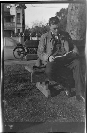 An unidentified man sitting on a wooden bench beneath a tree, reading and smoking a pipe, motorbike parked in street behind, large wooden building opposite, probably Christchurch