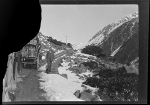 An unidentified man standing on the side of a mountain road in snow, next to horse-drawn coaches, Arthur's Pass, Selwyn District