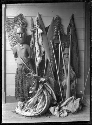 Maori carved figure wearing a piupiu and tiki, standing against a wall with weapons and cloaks.