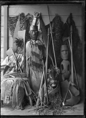 Maori carved figure wearing a piupiu and tiki, standing against a wall with weapons, implements and cloaks.