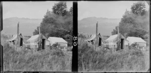 Unidentified man standing in front of huts made of canvas and plywood with his washing hanging near a wire netting fence, at a scheelite miners' camp, Paradise, Queenstown Lakes District