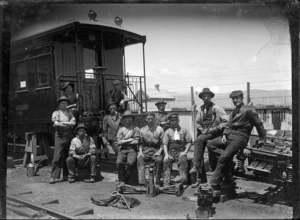 Group of railway workmen with various tools and implements, and the back of a passenger carriage visible.