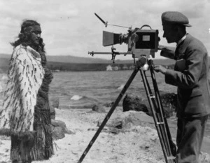 An unidentified man using a movie camera to film a Maori woman, standing by the sea, location unidentifed
