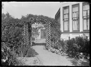 Part of a garden showing a trellis archway covered with climbing plants, with a view of a high trellis fence through the arch.