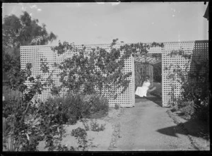 An unidentified woman sitting in a latticed arbour, seen through a gateway in a trellis fence partially covered with climbing plants.