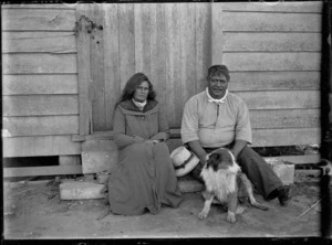 Unidentified Maori man and woman seated on a step in front of a door with a chain and padlock by the door handle