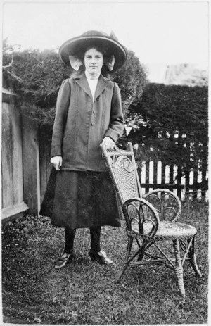 Unidentified young woman standing beside a cane chair in a garden
