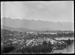 View of an unidentified settlement (possibly Queenstown) amongst trees beside a lake, with a mountainous range on the far side of the water.