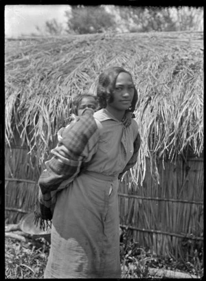 Young Maori woman carrying a child on her back, standing outside a whare thatched with palm fronds, at Rangiahua, 1918.