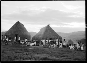 Group of local men, women and children, by thatched buildings, probably Fiji
