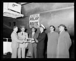 A group of men watch on as [Leo Lemuel White?] presents a copy of Whites pictorial reference of New Zealand to an unidentified pilot by air stairs of Pan American World Airways (Pan Am) plane with a sign for Polar Route flying from London and Paris on its fuselage
