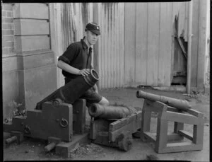 Unidentified schoolboy sitting beside 3 small military cannons [at a museum in Christchurch?]