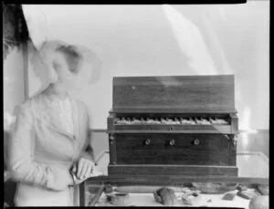 Unidentified woman next to a [museum?] display of objects including a small portable organ