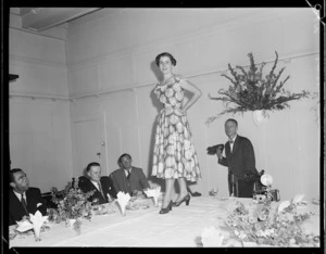 Model parading on table, men in suits seated, eating