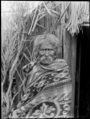 Maori man in a blanket outside a model raupo whare, at Christchurch Exhibition, 1906-1907