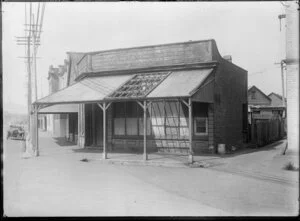 Shop on corner of unidentified street, apparently empty except for poster advertising Wood Milne shoe products in the window, probably Christchurch
