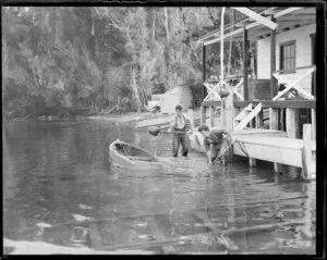 Two men in shallow water shoveling gravel into a small boat, beside a boatshed