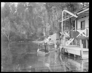Two men with shovels, one in a boat and one standing alongside in shallow water beside a boat shed