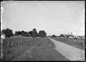 Country scene with a road running between two fields, and with two houses, one partially visible on the left, and a bungalow with verandahs on the right