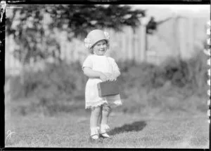 Toddler in hat and lacy dress and holding a box camera