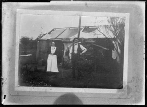 A hut with water tank on the left, and unidentified man and woman standing in front