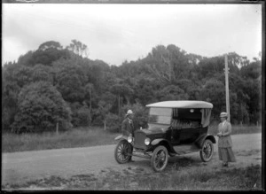 A man and woman standing beside a car on a road in a hilly, bushy area