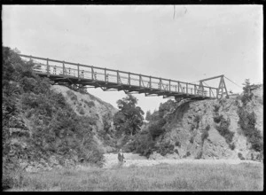 Woman (probably the photographer's wife Laura Godber) holding a billy while standing on the stony bank of a stream, beneath a suspension bridge